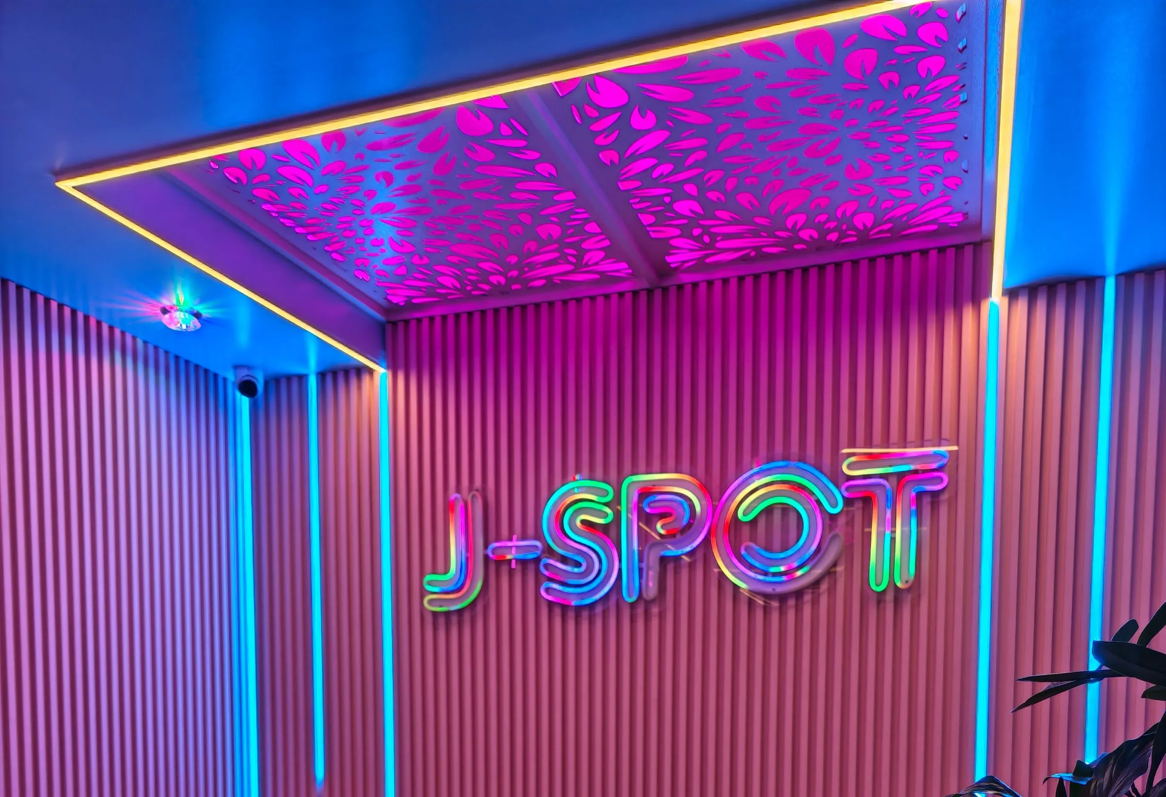 J-SPOT前台后方竖条纹墙上的彩虹色J-SPOT霓虹灯，两侧是青色灯带，上方是花纹天花板