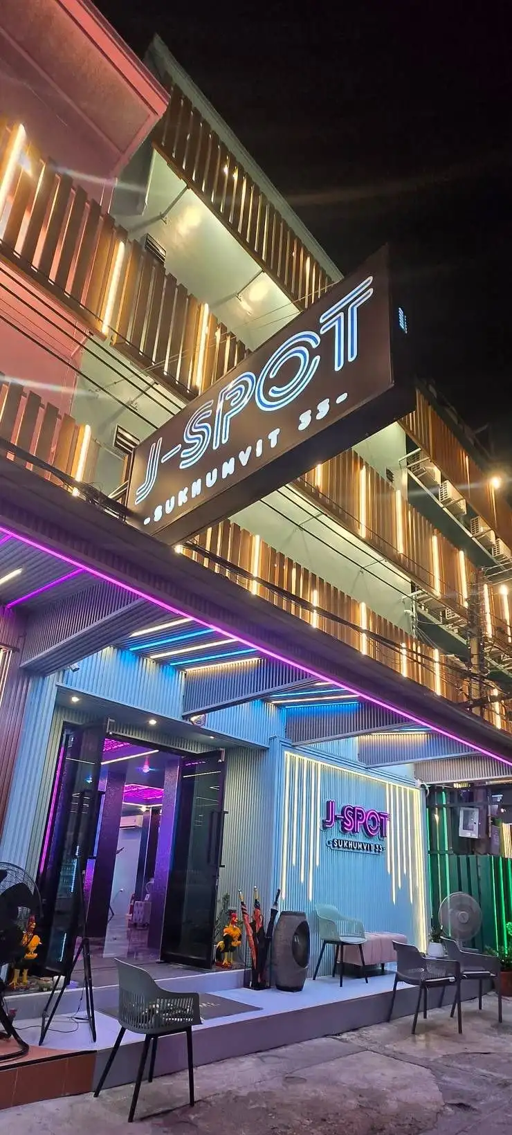 夜晚的J-SPOT：入口上方亮着的J-SPOT SUKHUMVIT 33竖招牌，紫色和青色的灯光雨棚，楼上亮着灯的木质阳台