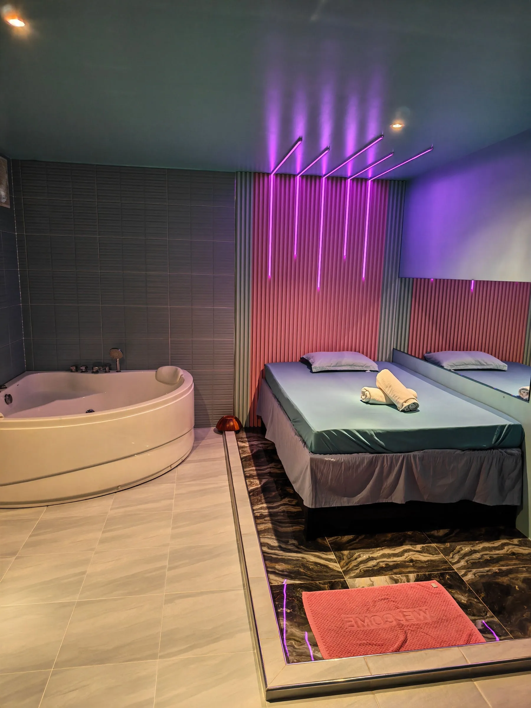 A private room at J-SPOT: a corner whirlpool bath, a massage bed with rolled towels, a long mirror and violet light lines on the wall