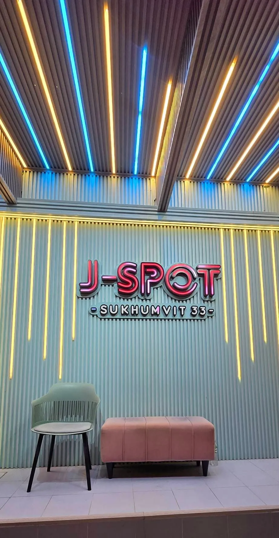 J-SPOTの外壁の「J-SPOT SUKHUMVIT 33」の看板。光のラインの下で灯り、前に椅子とベルベットのベンチ